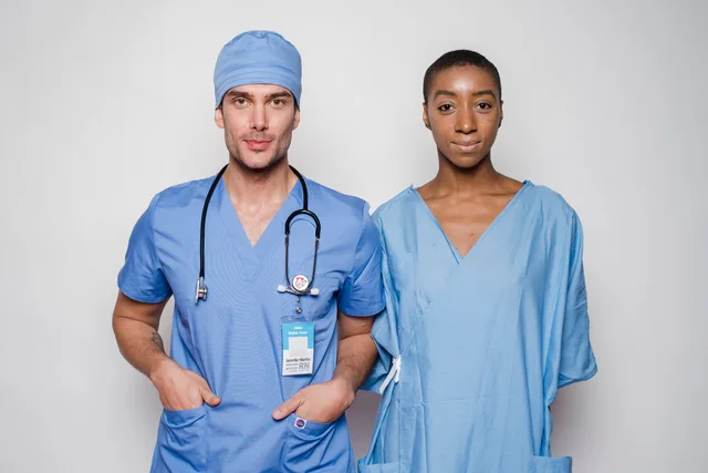 Positive male doctor in blue uniform with hands in pockets smiling and looking at camera while standing near nurse holding hands behind back in hospital. Autorisationskrav på tværs af danske brancher