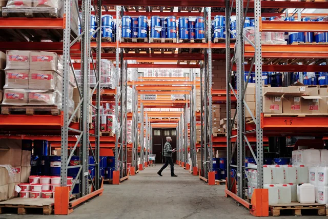 A man walking through a large industrial warehouse with stacked shelves filled with goods and products. Find de reelle ejere i CVR